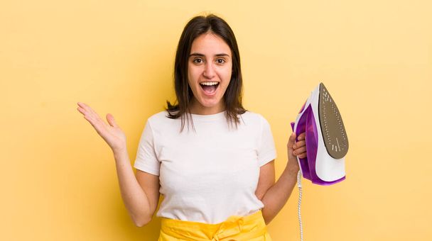 young hispanic woman feeling happy and astonished at something unbelievable. clothes iron concept - Photo, Image