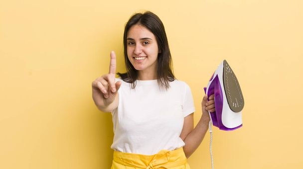 young hispanic woman smiling proudly and confidently making number one. clothes iron concept - Fotografie, Obrázek