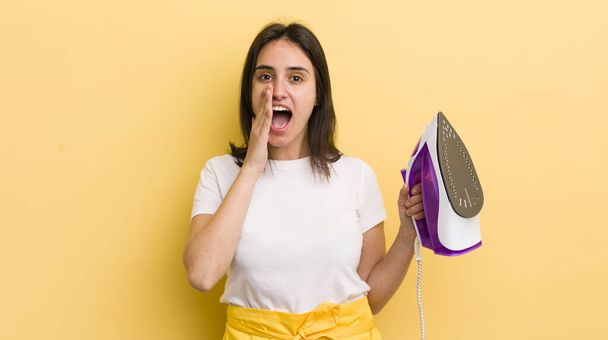 young hispanic woman feeling happy,giving a big shout out with hands next to mouth. clothes iron concept - Photo, Image
