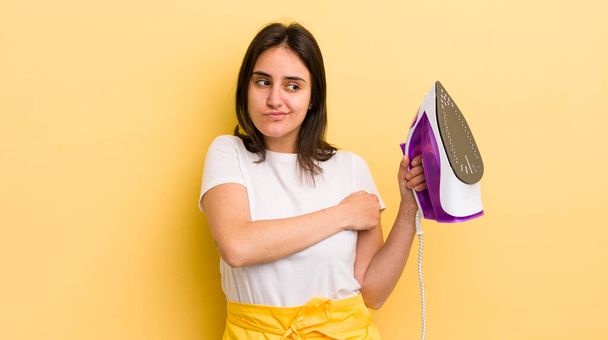 young hispanic woman shrugging, feeling confused and uncertain. clothes iron concept - Foto, afbeelding