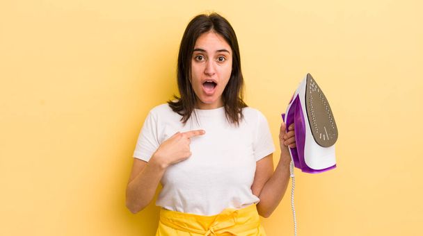 young hispanic woman looking shocked and surprised with mouth wide open, pointing to self. clothes iron concept - Foto, afbeelding