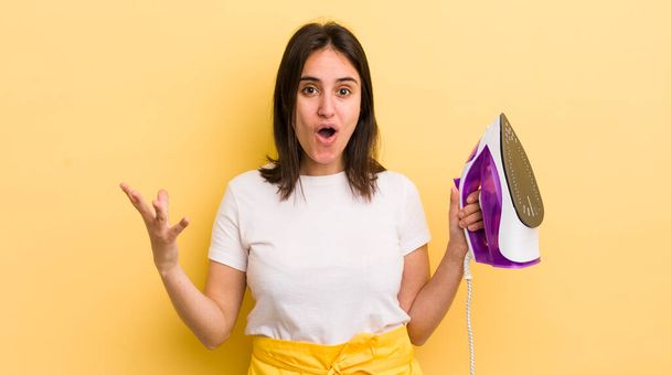 young hispanic woman feeling extremely shocked and surprised. clothes iron concept - Фото, изображение