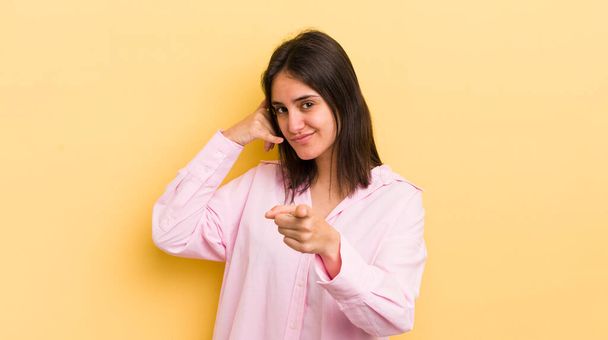young hispanic woman smiling cheerfully and pointing to camera while making a call you later gesture, talking on phone - Foto, imagen