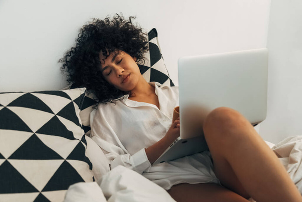 Молода афроамериканська жінка спить на ліжку з ноутбуком. Працюю пізно. - Фото, зображення