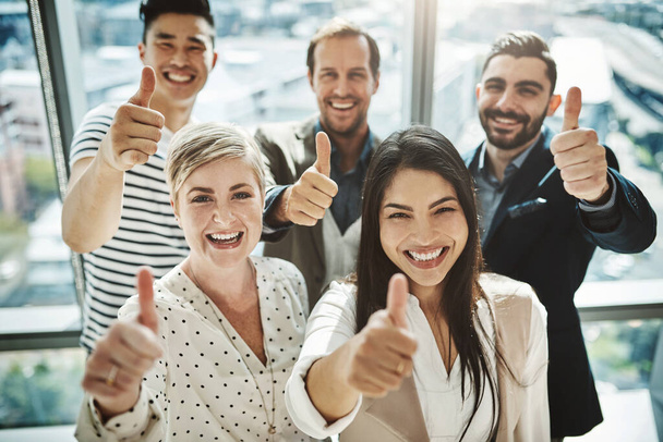 Their success rate is very high. Portrait of a group of cheerful businesspeople standing together while showing thumbs up to the camera inside of the office. - Foto, afbeelding