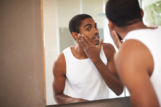 Er pflegt seine Haut. Ein junger Mann cremt sich sein Gesicht ein, während er in den Spiegel schaut. - Foto, Bild