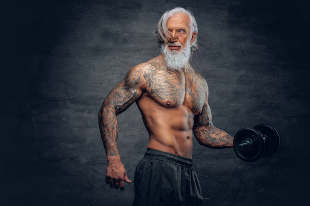 Bearded old man bodybuilder with tattoos holding dumbbell - Фото, изображение