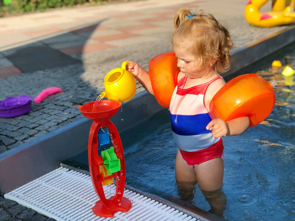 Niña preescolar salpicando en una piscina al aire libre en el cálido día de verano. Feliz niño sano disfrutando de un clima soleado en la piscina pública de la ciudad. Actividad infantil al aire libre con agua. - Foto, imagen