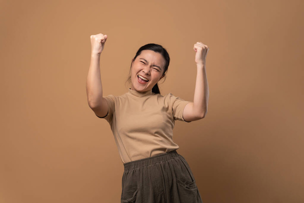 Azji kobieta szczęśliwy pewny siebie stojąc pokazując jej pięść zrobić zwycięski gest odizolowany na beżowym tle. - Zdjęcie, obraz