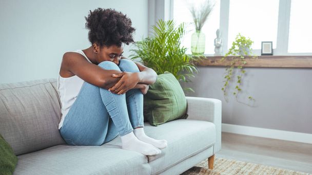 Young woman in gray clothes is sitting curled up on bed at home. Upset African American girl is suffering, crying. Depression because of loneliness, stress, problems. Psychological disorders concept. - Foto, Imagem