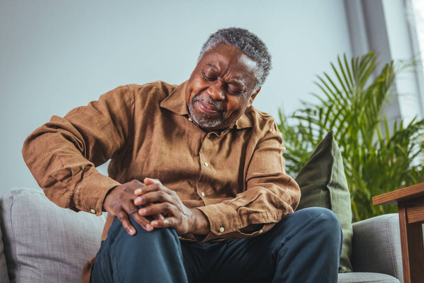 Photo of mature, elderly man sitting on a sofa in the living room at home and touching his knee by the pain during the day. Mature man massaging his painful knee. - Photo, image