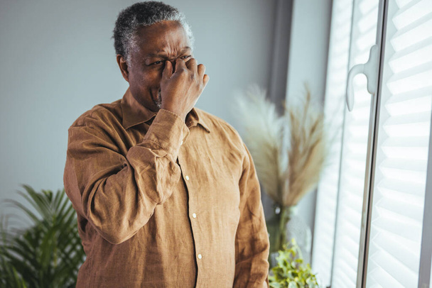A tired, depressed older African man is sitting on the couch in the living room, feeling hurt and lonely. An older, gray-haired man touches his head, suffers from a severe headache or remembers bad memories - Photo, Image