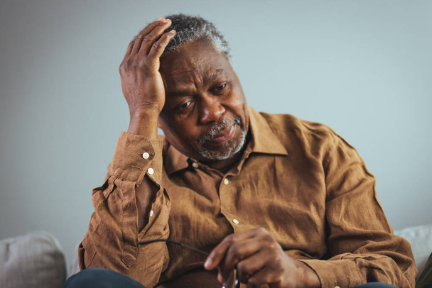 A headache. An elderly African-American who suffers from migraines and sits at home. Stressful older man sitting in bed at home alone, touching his head. Health care, health problems in old age concept. - Foto, imagen