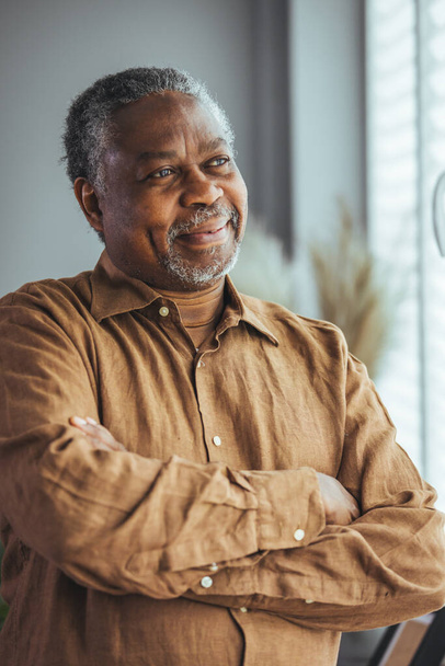 Side view of a happy African retired elderly man standing at home by the window. A satisfied old man looks out the window and smiles as he stands by the window. Positive and self-confident seniors enjoy retirement. - Zdjęcie, obraz