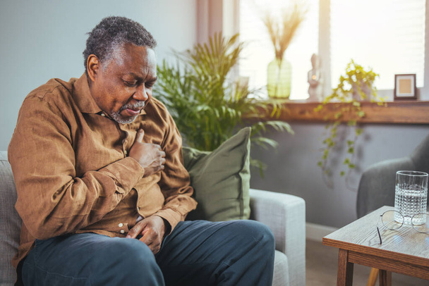 African senior man sitting on sofa at home with sickness. Elderly male suffering from heart attack and feel unwell need for medicine. Retirement people health care and illness recovery concept - Foto, Bild