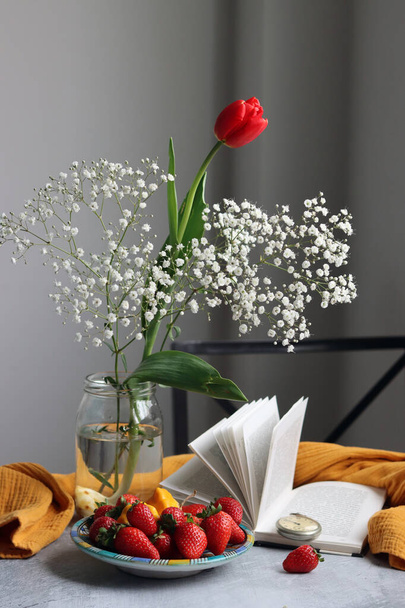 Tulipe rouge dans un bocal en verre, fraise sur une assiette, livre ouvert sur une table. Nature morte d'été avec des fruits.  - Photo, image