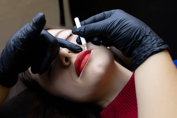 el procedimiento de tatuaje permanente del labio el maestro sostiene la punta de los labios con su dedo y aplica el contorno con un lápiz blanco - Foto, Imagen