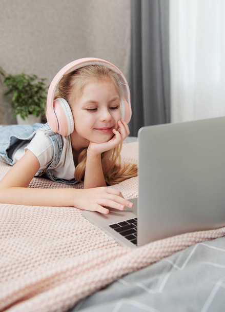 Посміхаючись щаслива маленька дівчинка, яка використовує ноутбук у бездротових навушниках у ліжку вдома
. - Фото, зображення