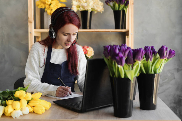 Młoda piękna kobieta kwiaciarnia w niebieskim fartuchu właściciela firmy za pomocą słuchawki i komputera podczas rozmowy z klientami online przyjmuje zamówienie online, zapisać długopisem w notebooku. - Zdjęcie, obraz