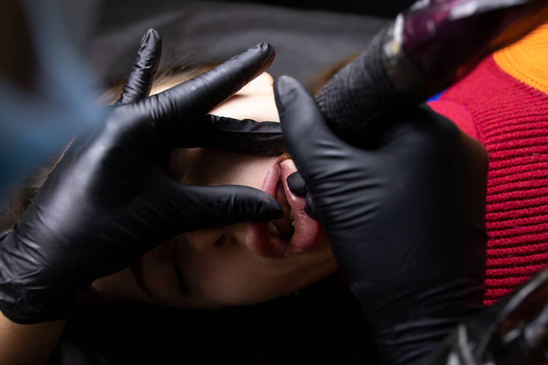 татуировщик в черных стерильных перчатках держит тату-машину и надевает татуировку на нижнюю губу модели - Фото, изображение