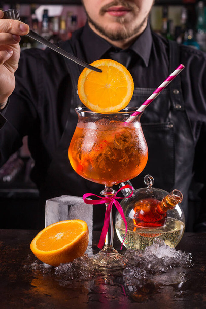 Bartender dressed in black apron and shirt is decorating orange cocktail drink on black background - 写真・画像