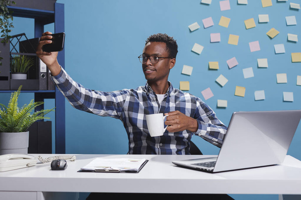 Σύμβουλος οικονομικής στρατηγικής λαμβάνοντας selfie φωτογραφία, ενώ πίνοντας ένα φλιτζάνι τσάι στο σύγχρονο γραφείο του χώρου εργασίας. - Φωτογραφία, εικόνα