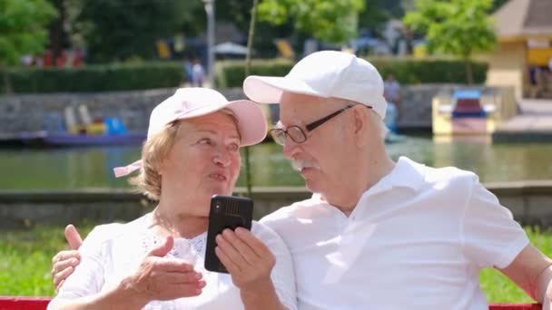 Een gepensioneerd stel dat op een bankje in het park zit en een nieuwe smartphone leert. - Video
