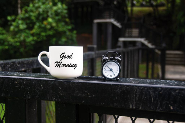 Guten Morgen Text auf Kaffeetasse mit Wecker und verschwommenem Park-Hintergrund. Morgenkonzept - Foto, Bild