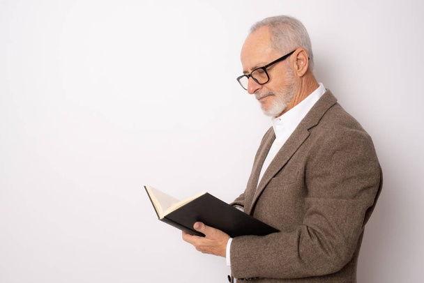 Retrato de homem sênior em roupas elegantes lendo um livro de pé isolado sobre fundo branco. - Fotografia, imagem