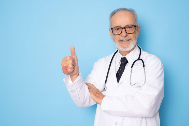 Senior grey-haired doctor man standing over isolated blue background happy face smiling with thumb up looking at the camera. Positive person. - Photo, image