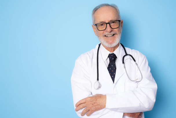 Senior grijs-harige arts man die staat over geïsoleerde blauwe achtergrond gelukkig gezicht glimlachend met armen gevouwen kijken naar de camera. Positief persoon. - Foto, afbeelding