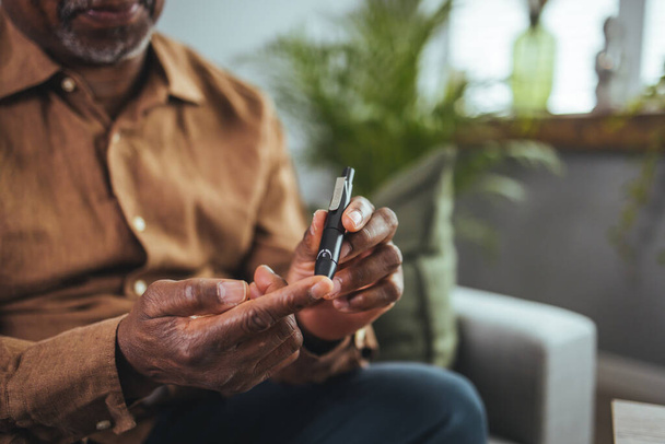 A modern African man sits at a table in the living room and takes blood from his finger due to diabetes. Everyday life of a man of African American nationality with a chronic disease who uses a glucose tester. - Foto, Imagem