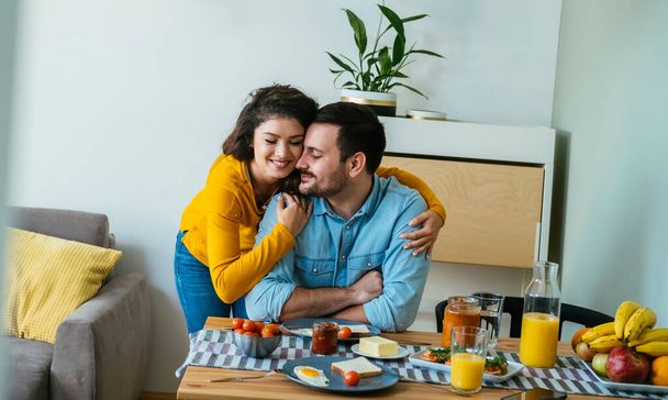 Gelukkig paar omhelzen tijdens het eten van ontbijt samen thuis. glimlachende vrouw knuffelen haar man terwijl het hebben van gezond ontbijt in de eetkamer. - Foto, afbeelding