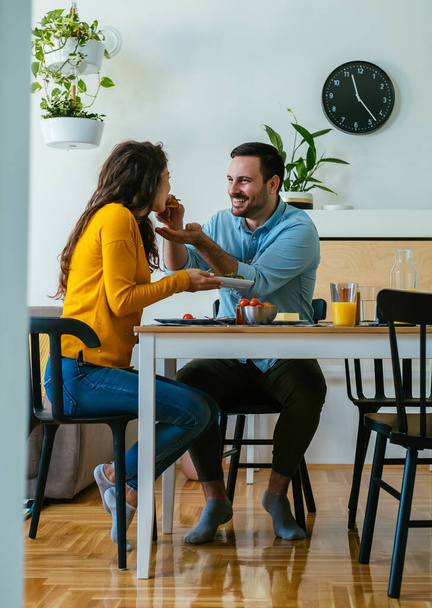 Счастливая пара завтракает вместе дома. Улыбающийся муж кормит жену и делится едой, сидя за столом в столовой. - Фото, изображение