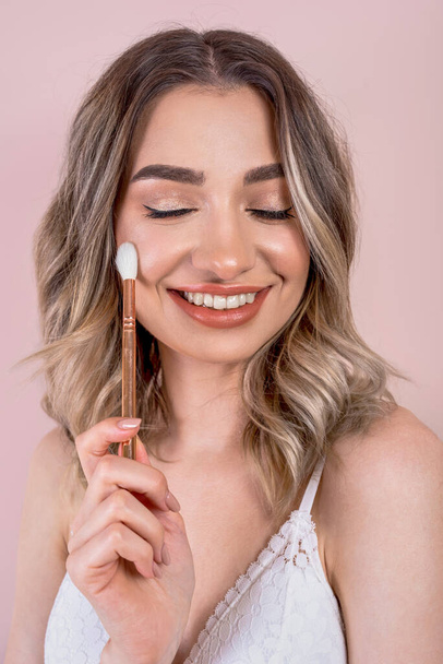 Beutiful blonde girl with natural makeup on light pink background, make up brushes in her hands, copy space - Foto, Imagen