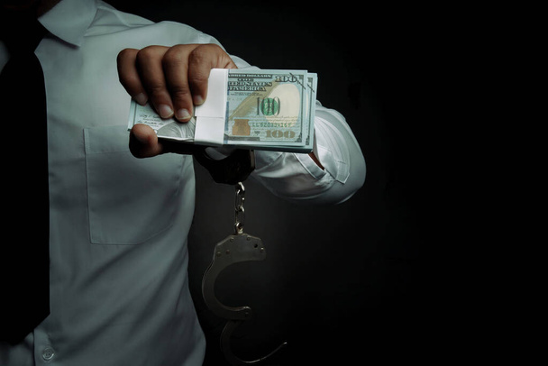 Νεαρός επιχειρηματίας στέκεται κρατώντας χρήματα και χειροπέδες που συνδέονται με το χέρι του Concept Διαφθορά της αστυνομίας, κακός μπάτσος, δωροδοκία της αστυνομίας. - Φωτογραφία, εικόνα