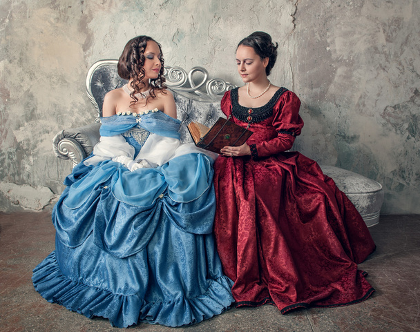 Two beautiful women in medieval dresses on the sofa reading book - Photo, Image