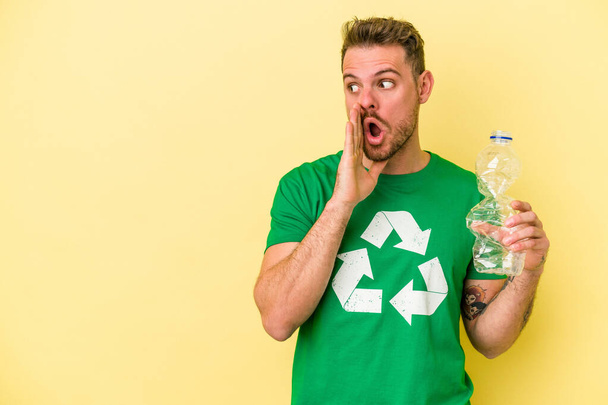 Молодий кавказький чоловік тримає пляшку пластику для переробки ізольованої на жовтому тлі каже таємні гарячі новини і дивиться вбік
 - Фото, зображення