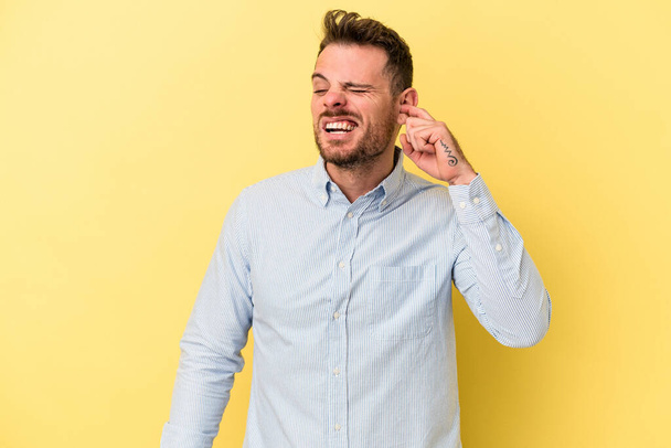 Young caucasian man isolated on yellow background covering ears with fingers, stressed and desperate by a loudly ambient. - Photo, image