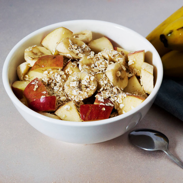 White bowl with chopped banana and apple with oatmeal a spoon on the side and part of ripe banana bunch on the side on a blue cloth - Photo, Image