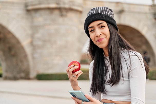 Funny hungry girl with appetite bites into a red apple sitting in the park looking at the camera with her smartphone. healthy life concept. copy space for text. - Foto, imagen