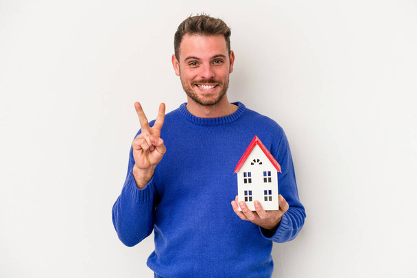 Young caucasian man holding a little house isolated on white background showing number two with fingers. - Foto, afbeelding