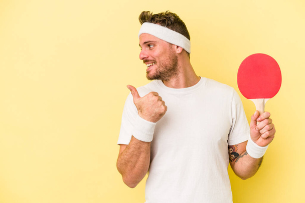 Junger kaukasischer Mann mit Tischtennisschläger isoliert auf gelbem Hintergrund, Daumenfinger weg, lachend und unbeschwert. - Foto, Bild