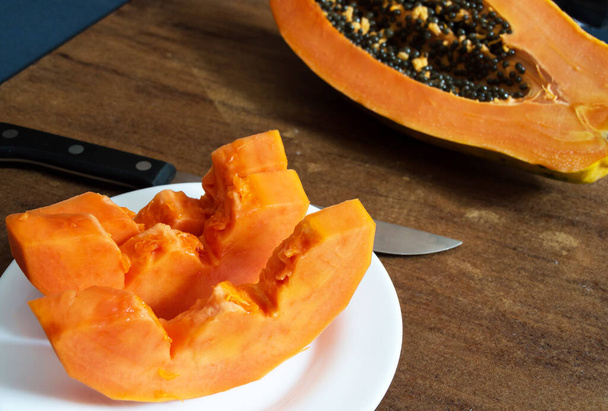 Three slices of papaya on a white plate with a knife on the left side of the image and part of a piece of papaya with seeds on the right of the slightly blurred image - Photo, Image