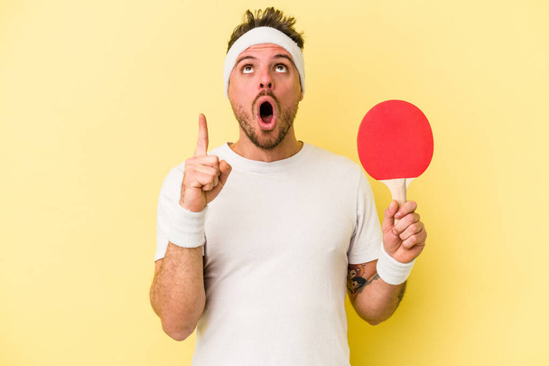 Giovane uomo caucasico tenendo racchetta da ping pong isolato su sfondo giallo che punta verso l'alto con bocca aperta. - Foto, immagini