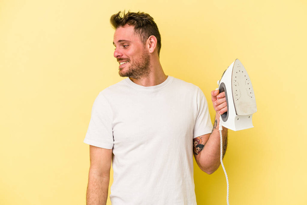 Young caucasian man holding a iron isolated on yellow background looks aside smiling, cheerful and pleasant. - Foto, Bild