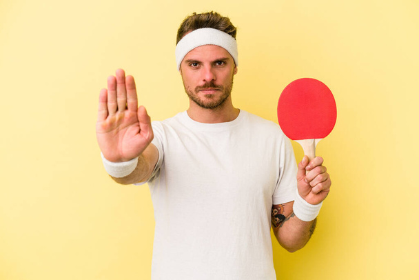 Jeune homme caucasien tenant raquette ping pong isolé sur fond jaune debout avec la main tendue montrant le signe d'arrêt, vous empêchant. - Photo, image