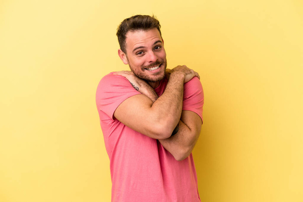 Young caucasian man isolated on yellow background hugs, smiling carefree and happy. - Fotó, kép