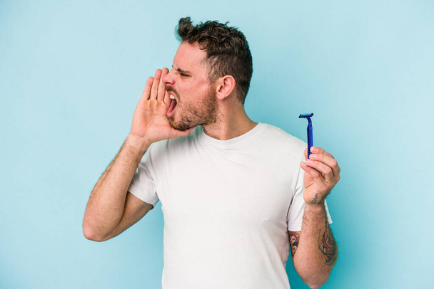 Giovane uomo caucasico radersi la barba isolato su sfondo blu gridando e tenendo palmo vicino bocca aperta. - Foto, immagini