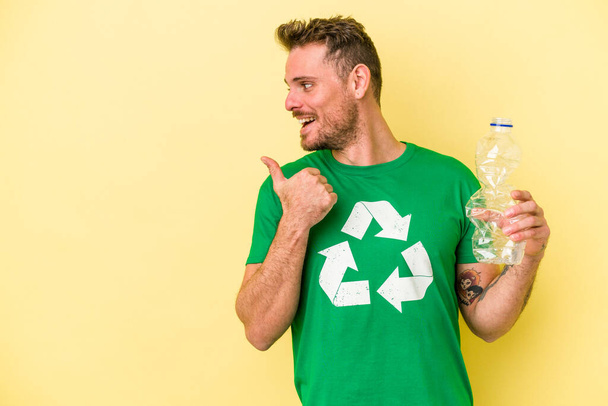 Jeune homme caucasien tenant une bouteille de plastique à recycler isolé sur des points de fond jaunes avec pouce loin, riant et insouciant. - Photo, image